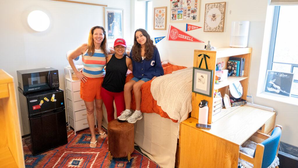 A new student sits on a dorm room bed and poses with two female relatives