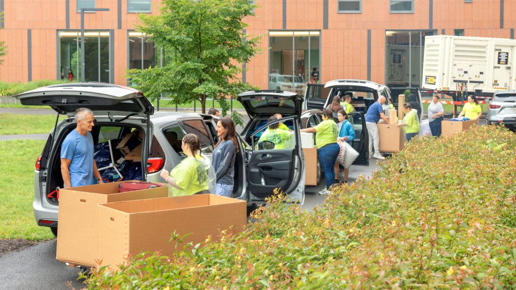 A line of cars with families unpacking belongings into bins with the help of movers in yellow shirts