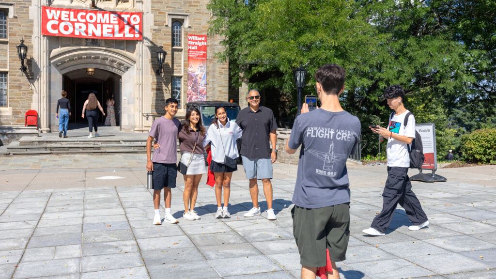 A family poses for a photo in front of the Straight with a WELCOME TO CORNELL sign