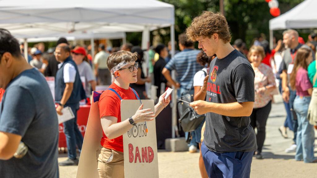 a student wearing a "Join Band" placard chats with another student on Ho Plaza during Move-In events, August 2026