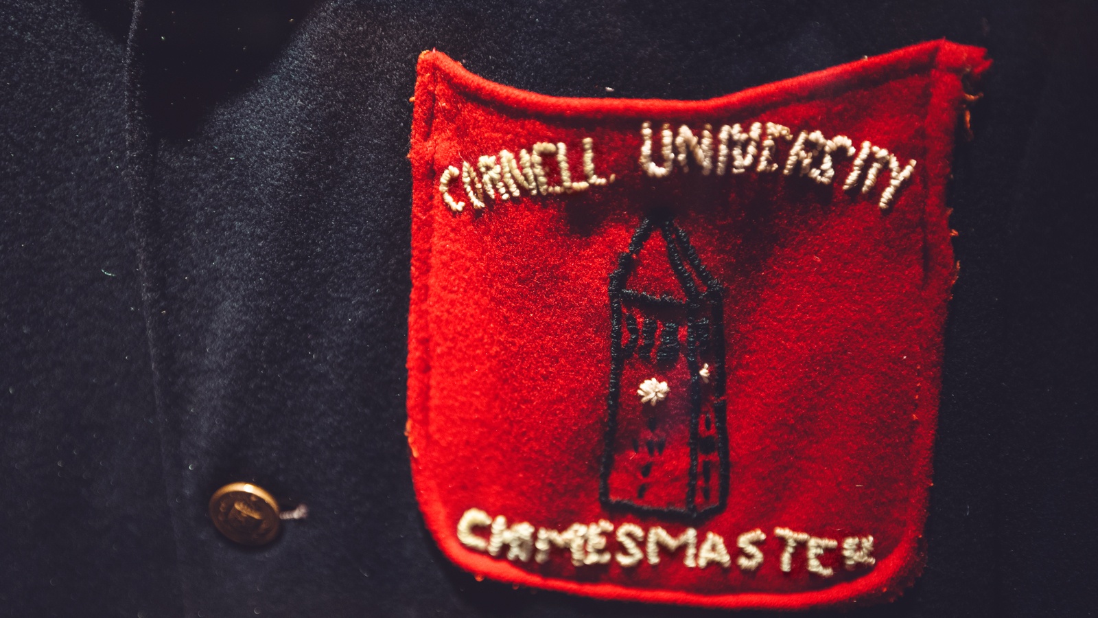 Detail view of a chimesmaster jacket, showing a red pocket embroidered with the words "Cornell University Chimesmaster" and a small rendering of McGraw Tower; the jacket was donated by Paul Lyon ’64 and is on display in the Chimes Museum