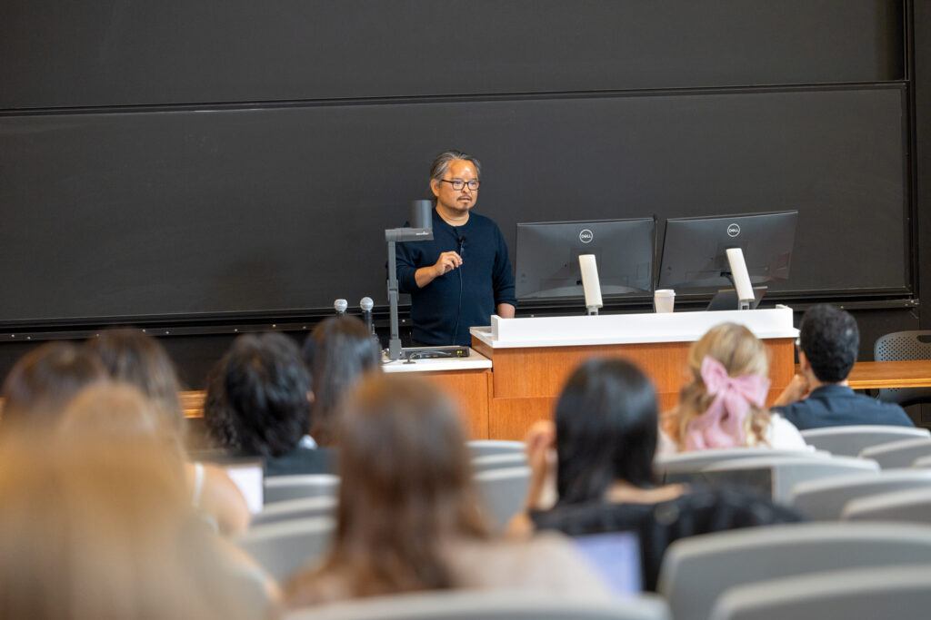 psychology professor Anthony Ong stands at the front of a classroom next to two computer monitors while teaching The Science of Well-Being