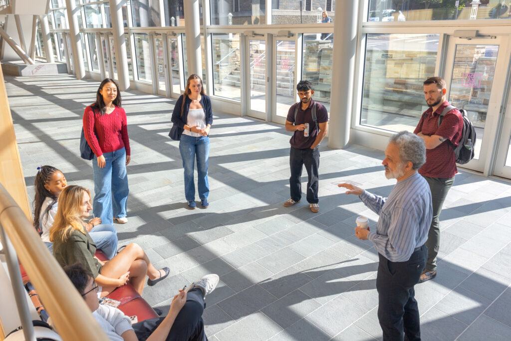 A group of seven students standing and sitting in a circle in the Beck Center atrium at Statler Hall while listening to a professor speak with a coffee in his hand