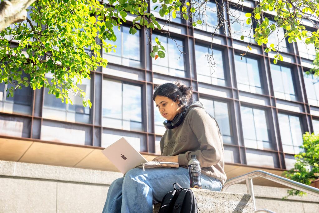 A student wearing jeans and a sweatshirt sits outside of Uris Hall and looks at her laptop