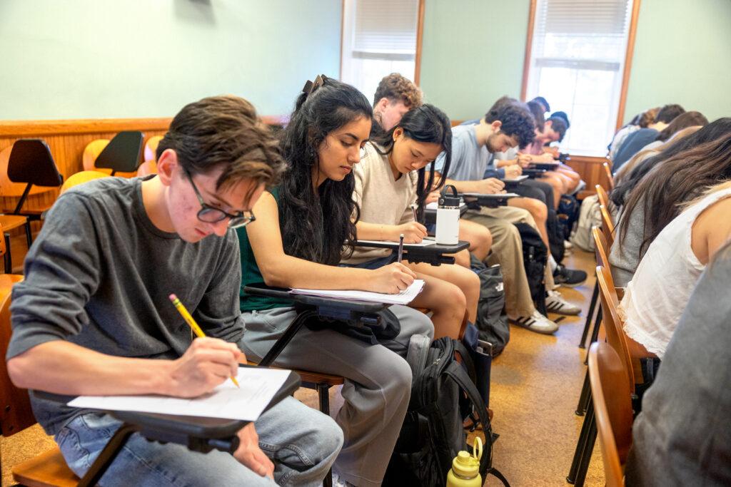 Two rows of students in a classroom, all focused on writing on papers in front of them