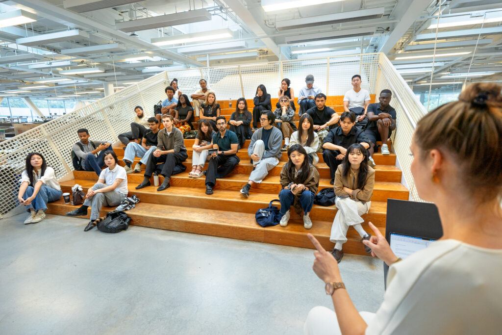 Students sit in a classroom on a set of broad steps and listen to a lecturer in front of them