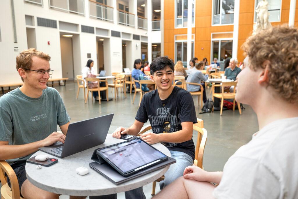 Three students sit at a table together while talking and laughing in the Klarman Hall atrium