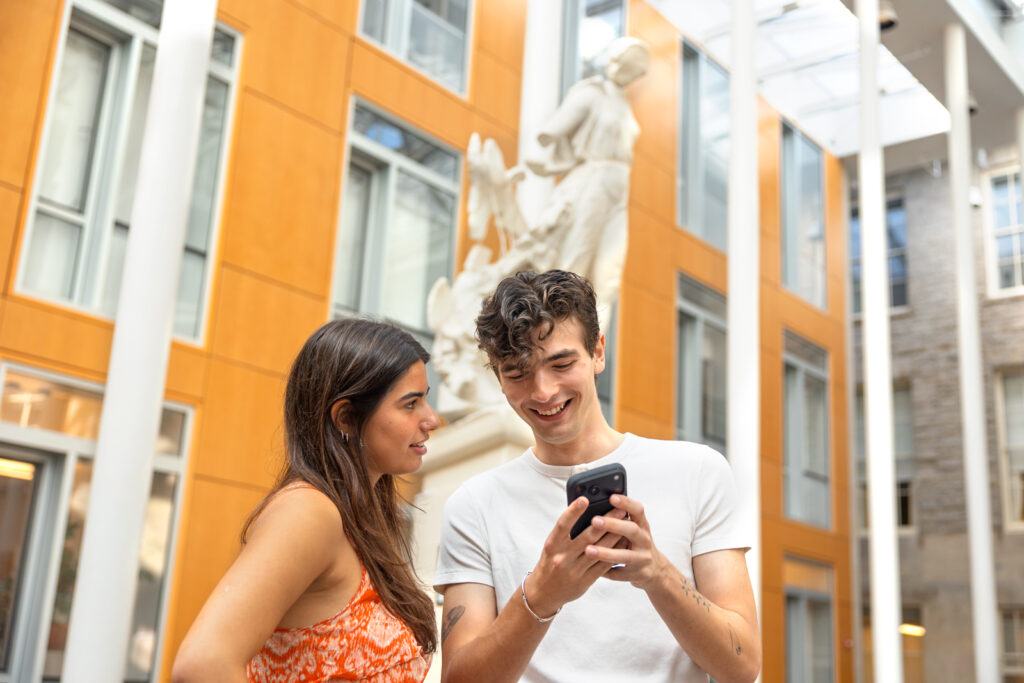 Two students look at a phone screen and smile in the atrium of Klarman Hall