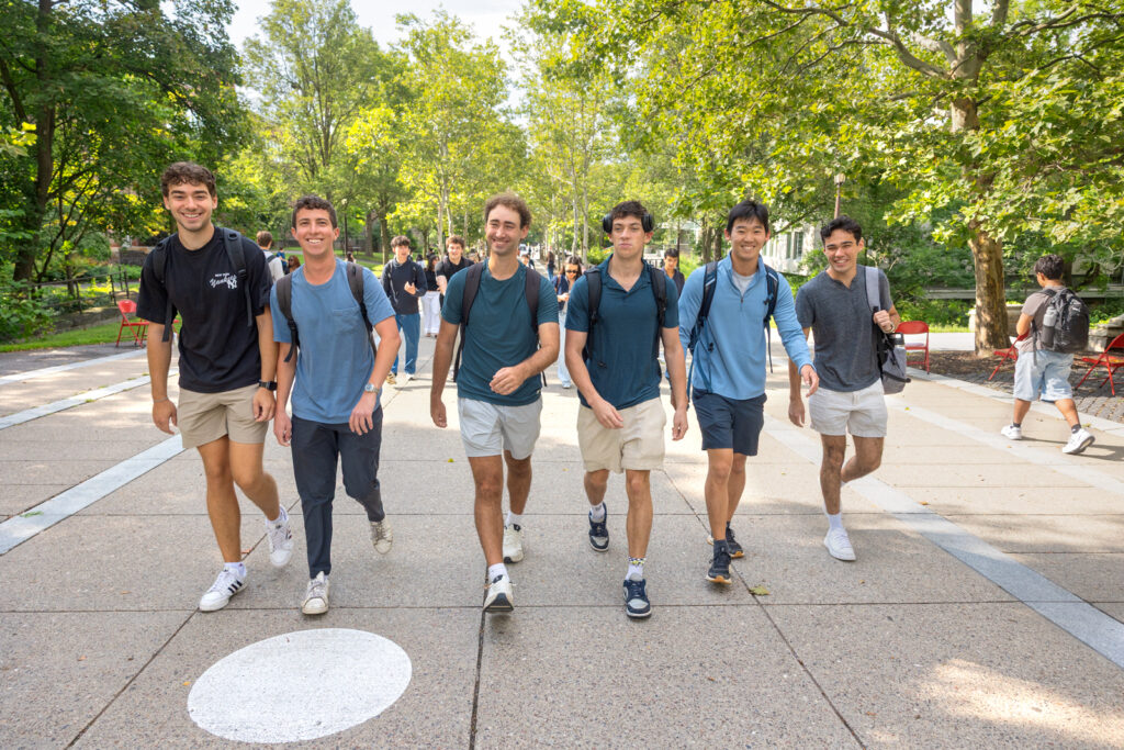 A group of six students in a line smile and walk together through Ho Plaza on campus