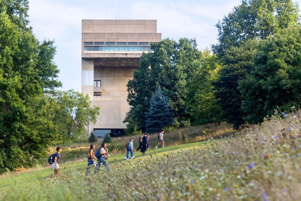 Students walk up the Slope on campus with their backpacks, with the Johnson Museum in the background and wildflowers in the foreground