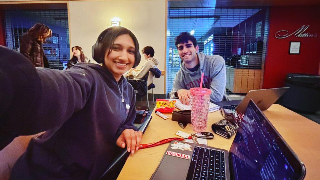 Nour Gajial and Yanni Kouloumbis sit at a table at Cornell together wearing sweatshirts and work on MathGPT on their laptops.