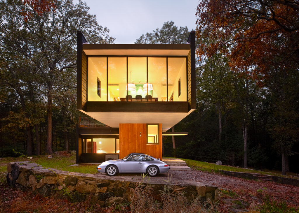Photo by Brad Feinknopf ’86 of a private residence, with an illuminated upper floor and a cantilevered space with a car parked underneath, surrounded by woods and a stone wall, in Waccabuc, NY