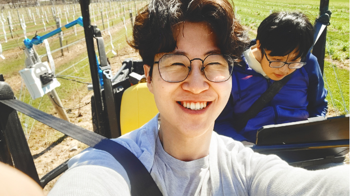 Jonathan Moon takes a selfie while working in the field with Ertai Liu behind him looking at a laptop.