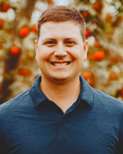 Bobby Brown in a navy blue collared shirt smiling in front of an apple tree.