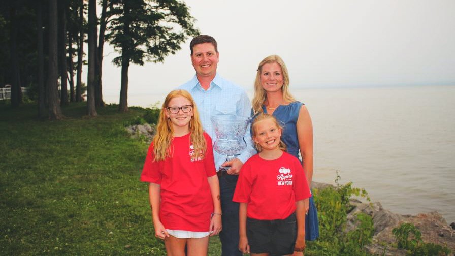 Bobby Brown holds the Apple Grower of the Year trophy with his wife and two daughters around him next to a lake.