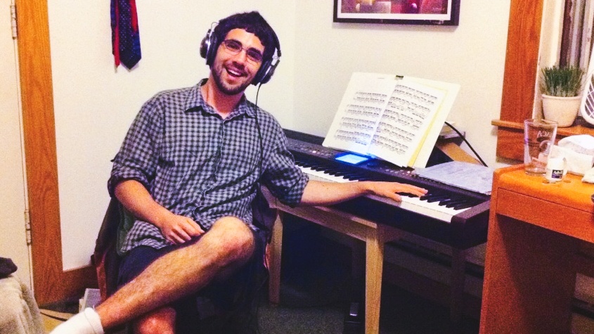 Daniel Anastasio as a student in his dorm room smiling with his hand on a keyboard.