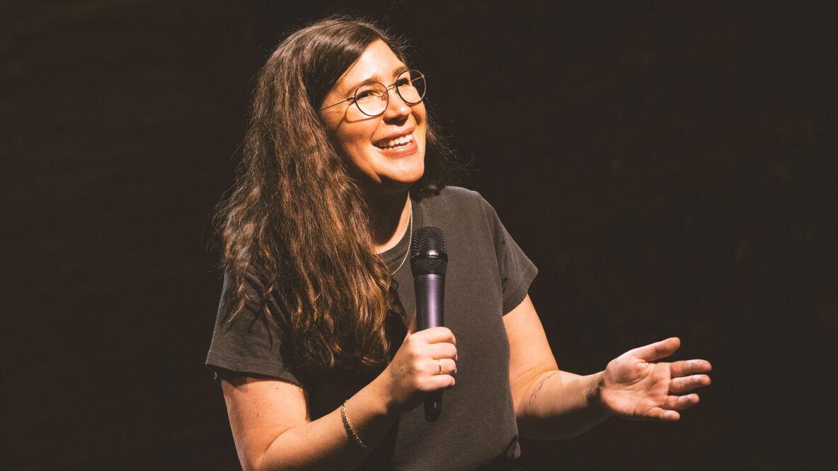 Alison Leiby in a black T shirt and microphone in hand during a performance of Oh God: A Show About Abortion