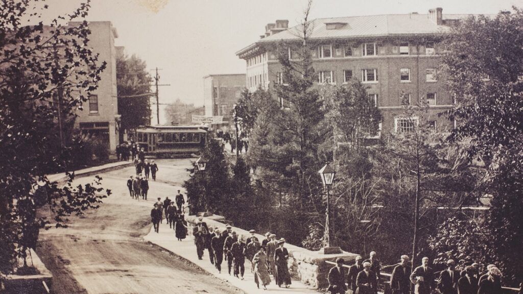 pedestrians cross the bridge in Collegetown to campus, with a trolley car and Sheldon Court in the background, early 1900s