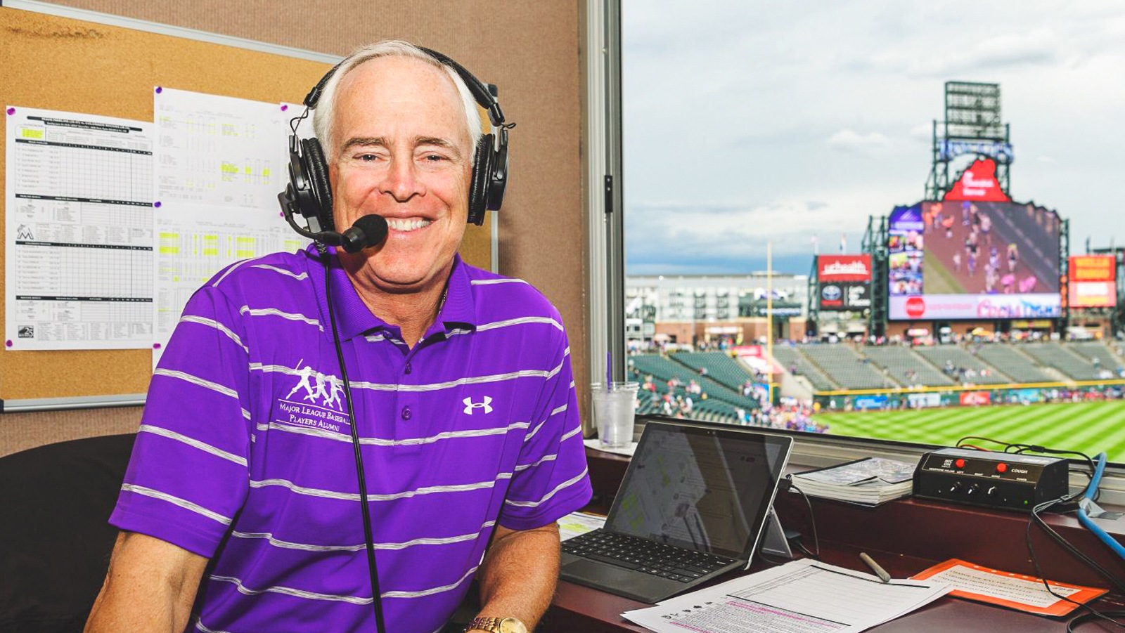 Corrigan smiles from the broadcast booth at Coors Field with the baseball field behind him.