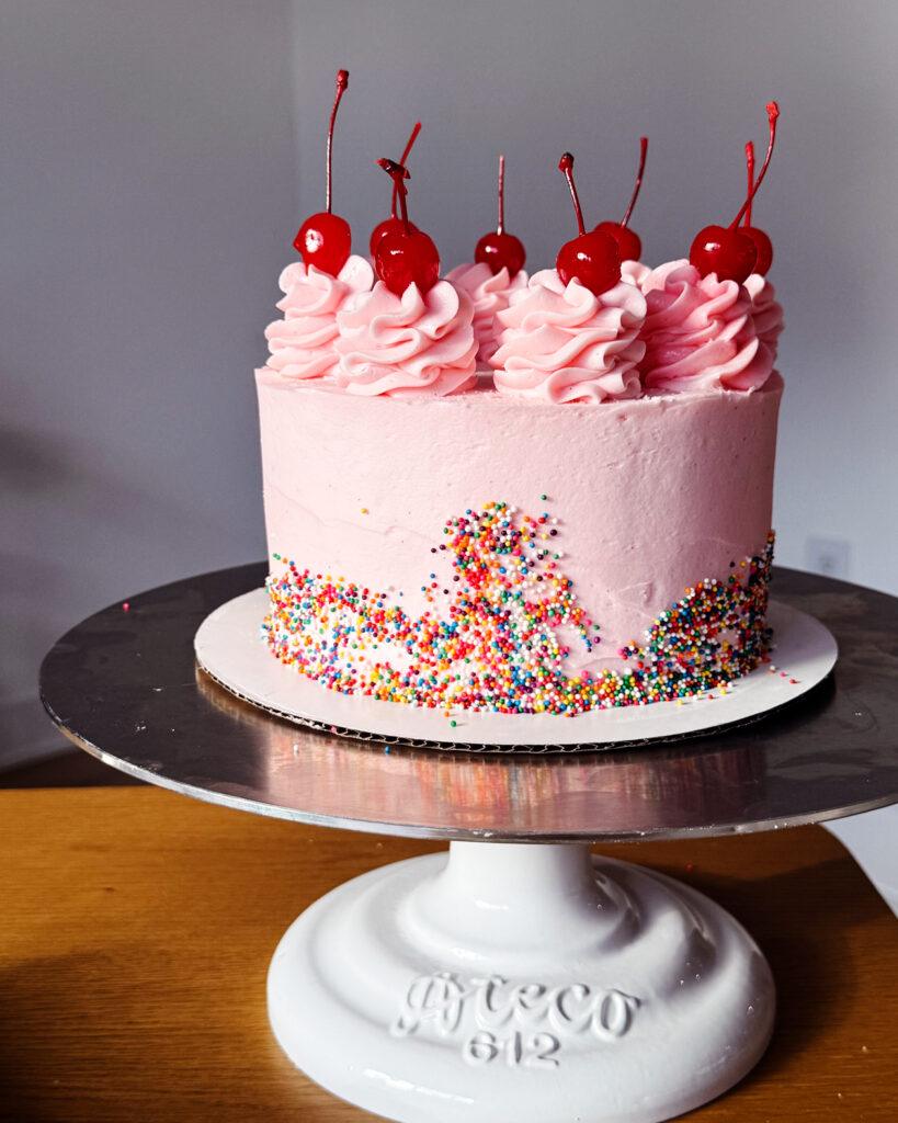 A cake on a stand with rainbow sprinkles, pink icing and cherries