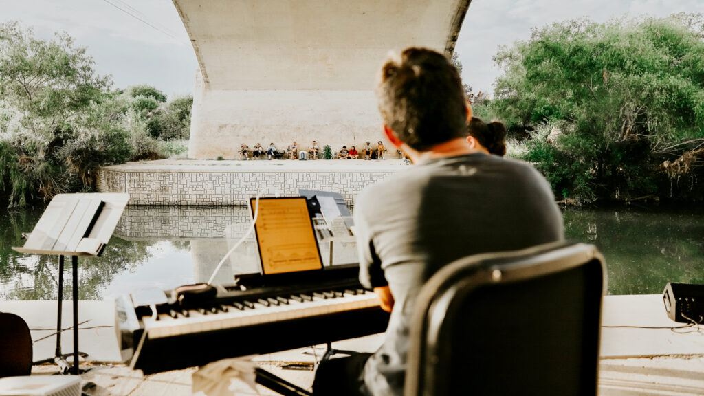 Daniel Anastasio plays piano outdoors to a group listening on the other side of a canal.