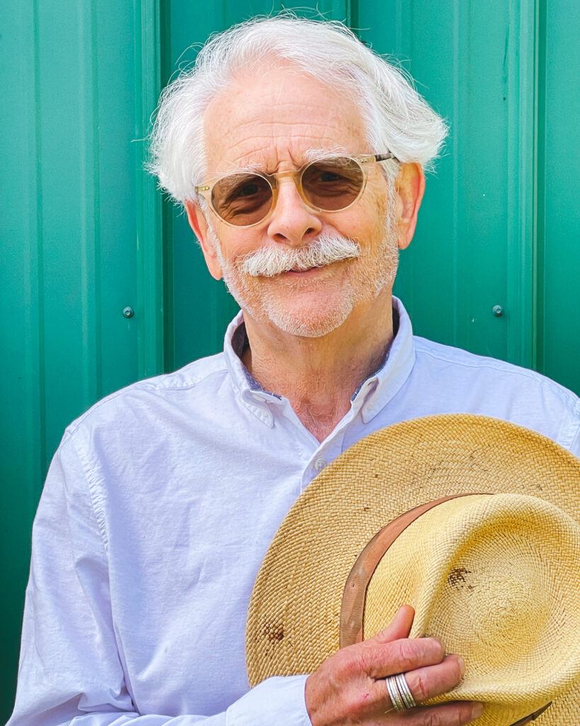 Kent Diebolt smiles wearing glasses and a white dress shirt while holding a tan, woven hat in his hand.