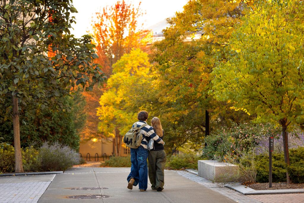 Two students walk with their arms around each other on a campus pathway lined with trees with green, yellow, and orange leaves.