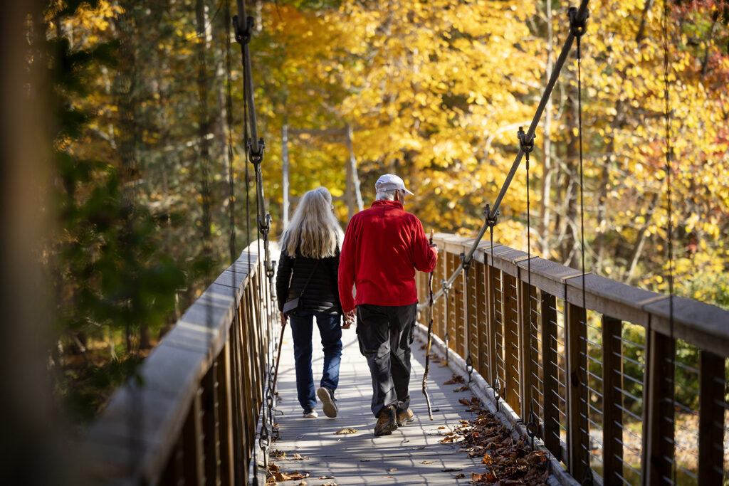 A man and a woman walk across the bridge over Flat Rock at Six Mile Creek and look at the trees.