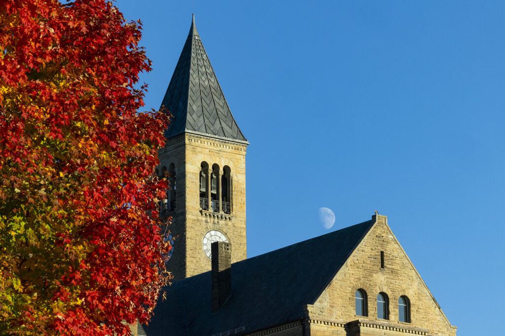 The moon is seen in a daytime blue sky above McGraw Tower and Uris Library with a red and green tree visible on the side of the building.