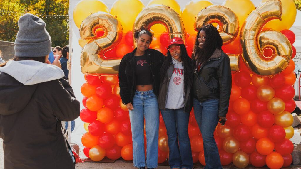 Three students pose for a photo in front of the balloon wall that says "2026" during fall-themed events on Ho Plaza at Homecoming