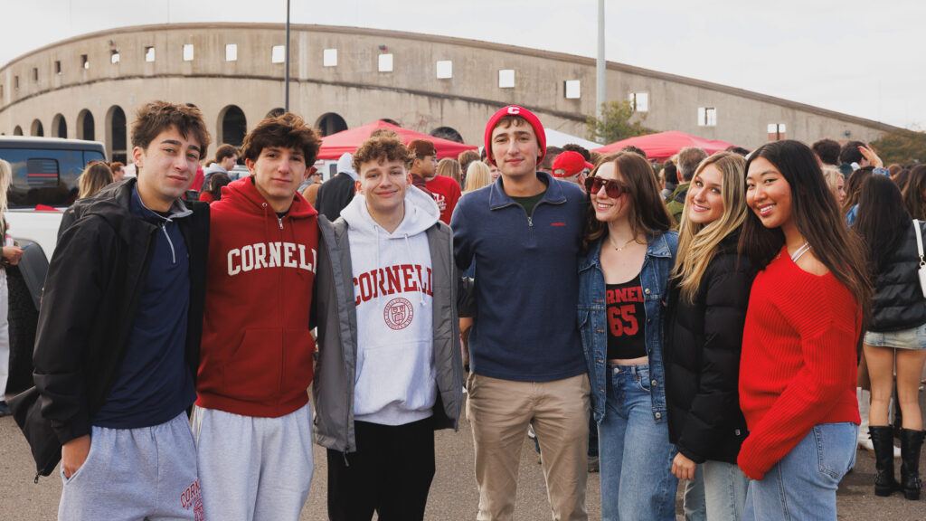 Seven Cornell students in red, white, and navy clothes smile for photo together in front of the crescent.