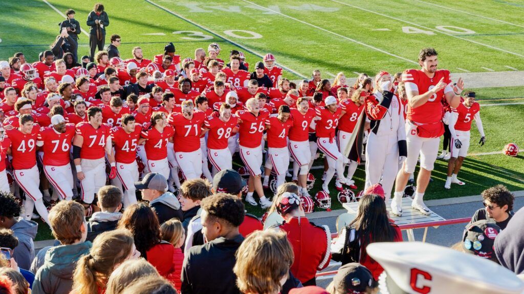 The Cornell football team stands on the field and faces fans with their arms around each other.