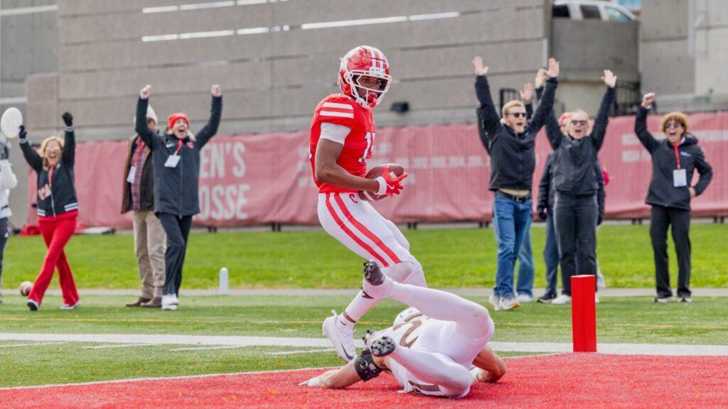 A Cornell player catches the ball in the end zone while spectators in the back hold their hands up to signal a touchdown.