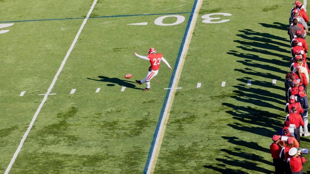 A Cornell football player kicks the ball as a row of spectators wearing red watch from the side.