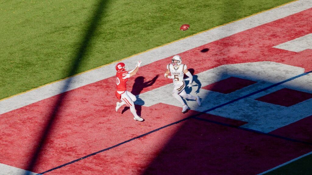 A Cornell football player holds his hands out to catch the football in the end zone while a Lehigh player runs towards him.