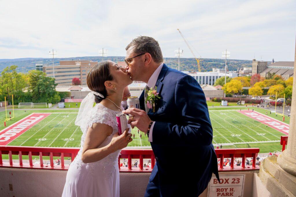 Yuko Jingu and Jeffry Clausen Jr. kiss with beer cans in their hands from their box overlooking the football game.