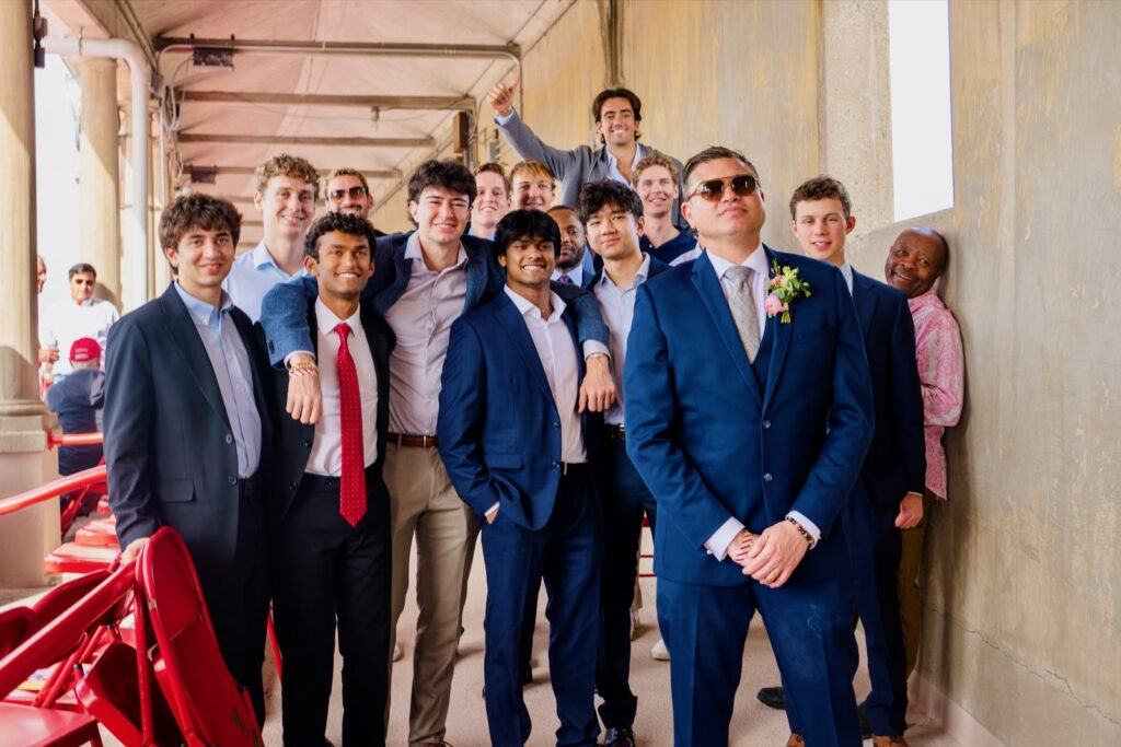 Jeffry Clausen Jr. smiles for a photo with students on the rowing team in suits behind him.