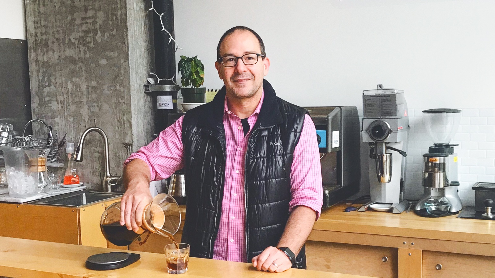 Matthew Berk pouring coffee at a counter, wearing a black vest and pink button-down shirt