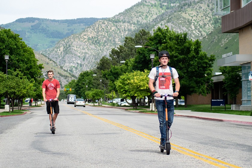 Two young men riding Spin electric scooters down a city street backed by mountains.