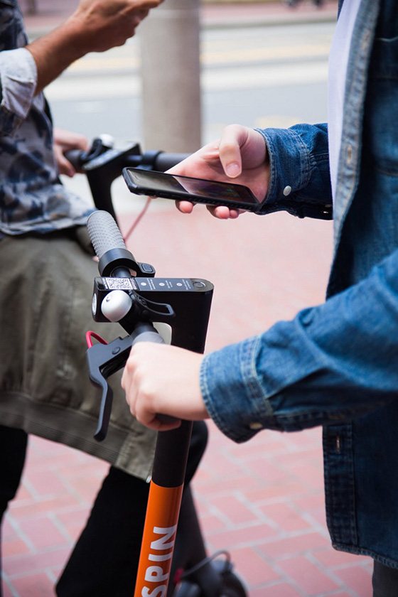 A young person holds a mobile phone in one hand and a scooter handle in the other.