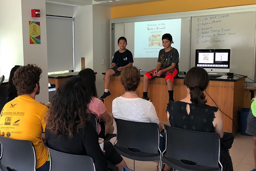 Two boys sitting on a lab table, talking to a small audience.