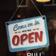 A sign reading "Come on in, we're open" hangs above white text that states "Pull handle" on a glass door