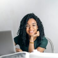 A young Black woman smiles toward the camera while sitting at a desk with a laptop. Her chin is resting on her palm.