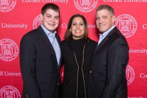 Lorette, her son and husband pose in front of a red and white Cornell background. 