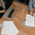 Two people sit at a table covered with papers, a laptop, and glasses of water