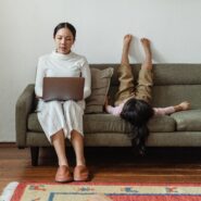 A mother works on a laptop while her child lays upside down on the couch next to her