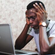 A man sits in front of a laptop with hands on his head, looking frustrated