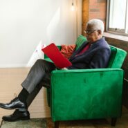 An older Black man sits on a couch with a folder in his hands.
