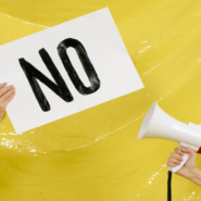 Two people holding a sign that says "No" and a mega phone in front of a yellow background