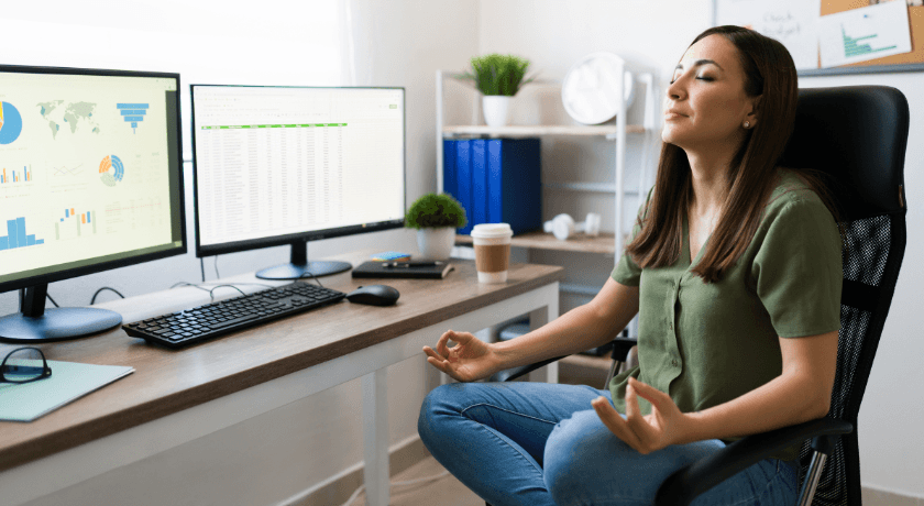 A woman sitting cross legged in an office chair, eyes closed and hands held in a meditation pose